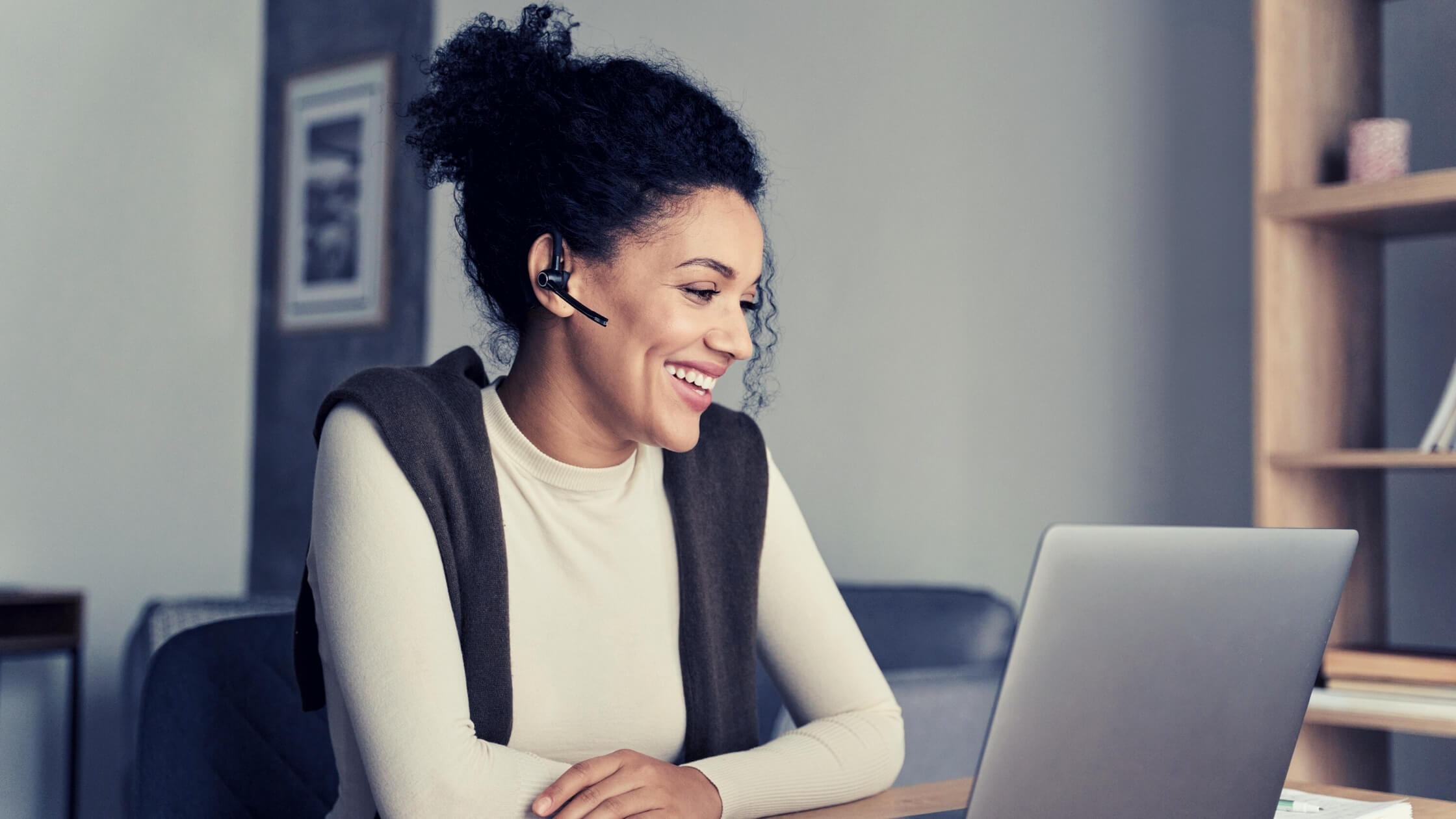 Mujer con el cabello recogido y un dispositivo de manos libres, de unos 40 años, sonriendo mientras mira la pantalla de su laptop
