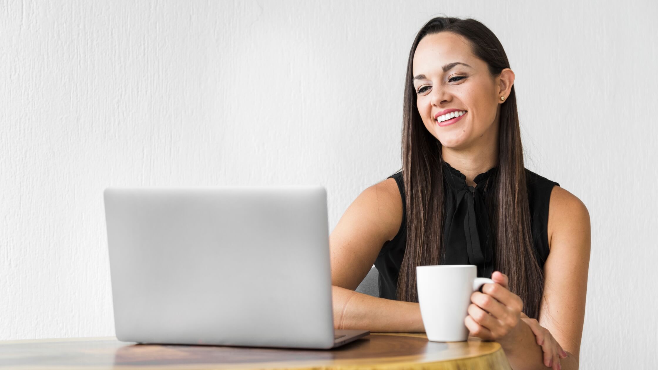 Muier vestida con blusa color negro, sonriendo mientras mira su laptop y sostiene una taza blanca de café en su mano